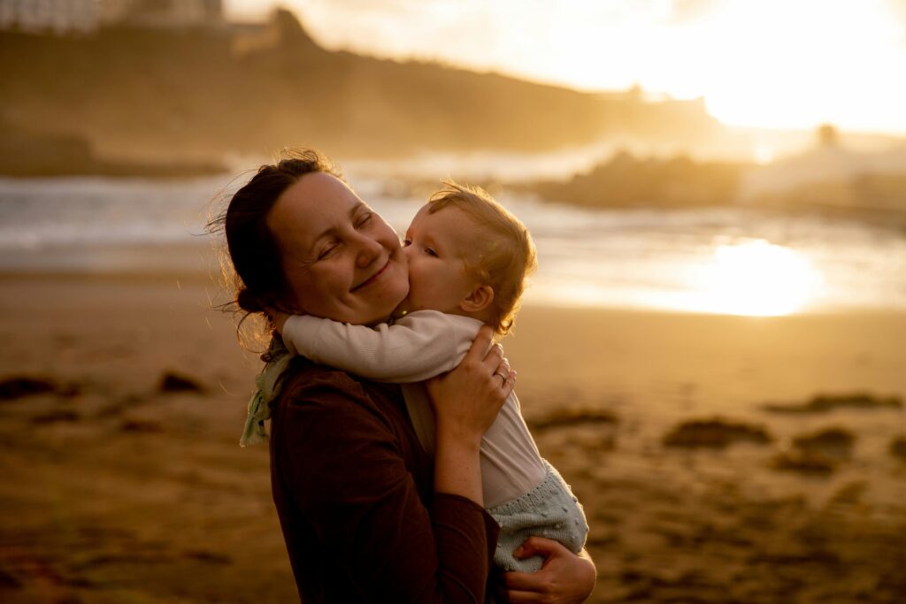 Uma mãe segura uma criança pequena no colo que  lhe abraça e beija. A mãe está de olhos fechados , sorriso largo em plena satisfação. Ao fundo a imagem desfocada de uma praia