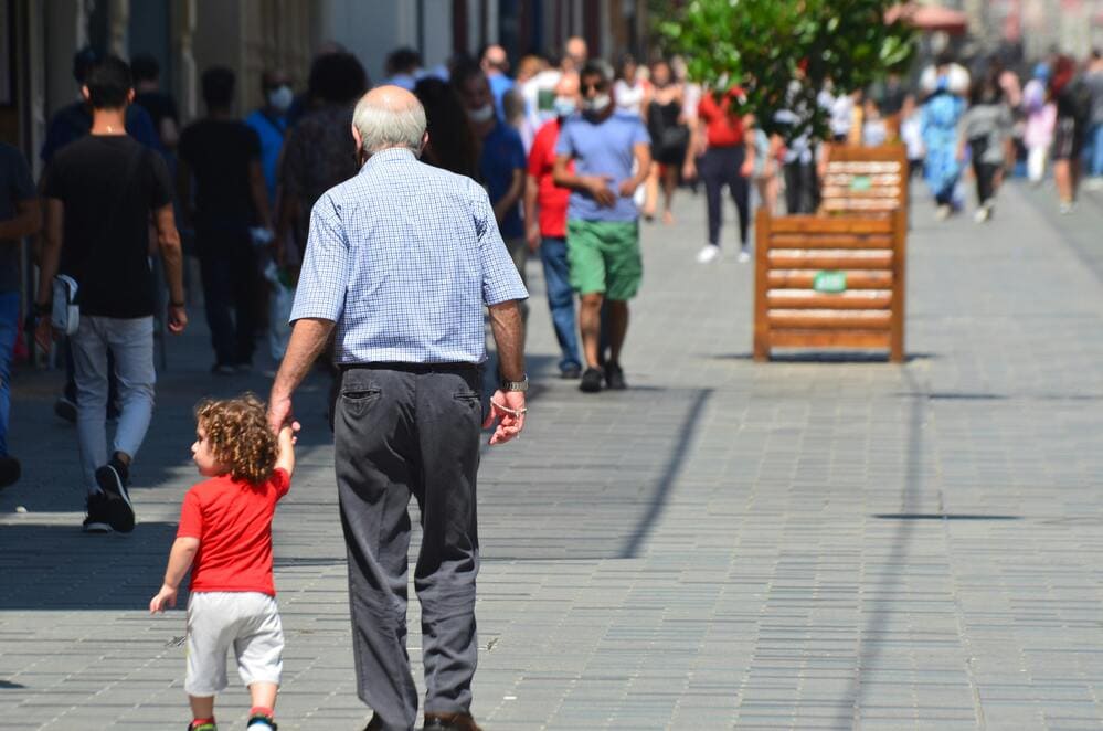 Um senhor de cabelos brancos caminha de mãos dadas com uma criança pequena em meio a cidade agitada